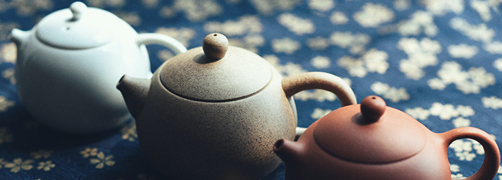 3 ceramic teapots in a row, in different shades of brown and grey, on a blue tablecloth with white flower detail